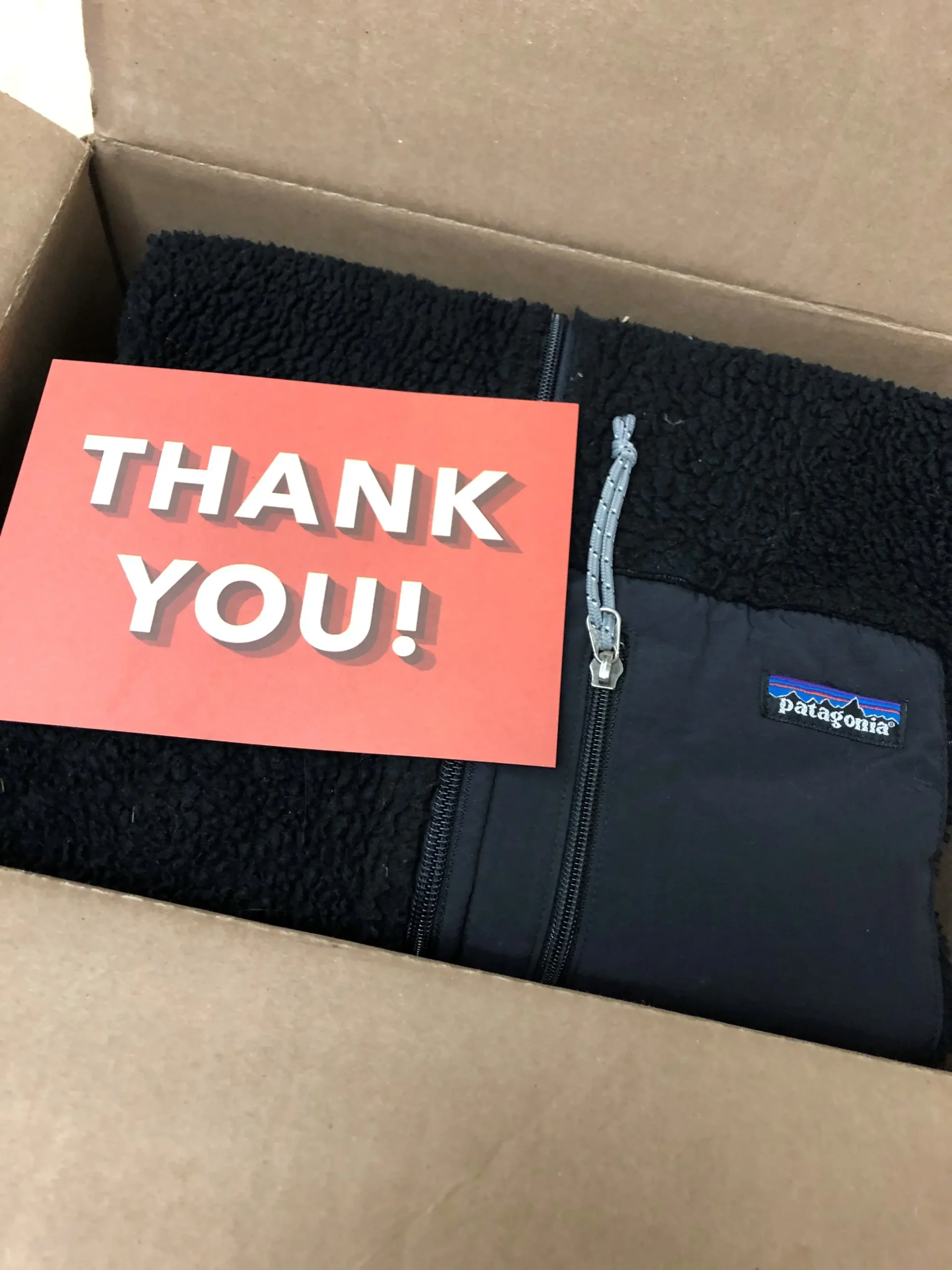 An open shipping box containing a black Patagonia fleece jacket accompanied by a vibrant "THANK YOU!" card